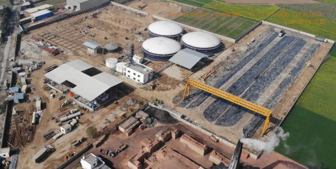 Aerial view of the Yamunanagar compressed biogas plant under construction, showing three digester domes and covered substrate windrows